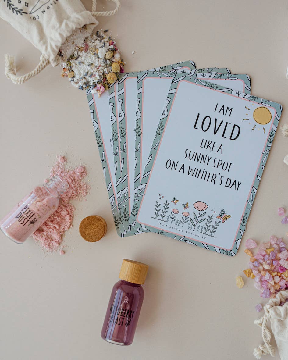 A set of botanical-themed affirmation cards reads, "I am loved like a sunny spot on a winter’s day." Surrounding the cards is THE LITTLE POTION CO's Enchanted Garden MINDFUL Potion Kit, which includes small bottles of colored powder, mixed dried flowers, and assorted small crystals.