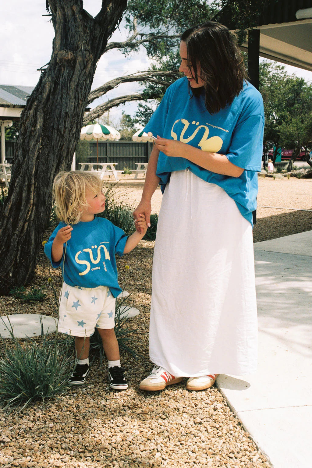 A woman and child stand outdoors by a tree in blue shirts with yellow designs. The woman points at the child while holding hands. The kid wears SUNDAY SIBLINGS' Sunday Siblings ~ Kiddo Shorts Blue, featuring an elastic waistband. They stand on a gravel path amid some greenery.