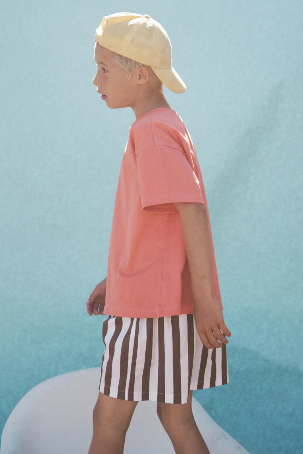 A child in a yellow cap, pink t-shirt, and LITTLE THE LABEL's Tei Shorts Brown/White Stripe walks past a blue background.