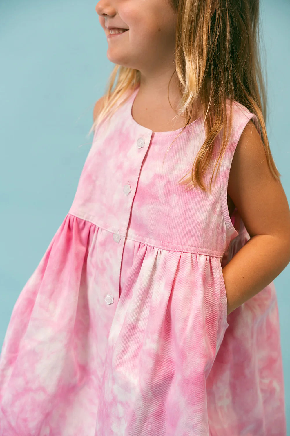 A young girl in a relaxed-fit Lollipop Dress by LENN LABEL, featuring a pink and white tie-dye pattern, stands against a light blue background, smiling slightly to the side.