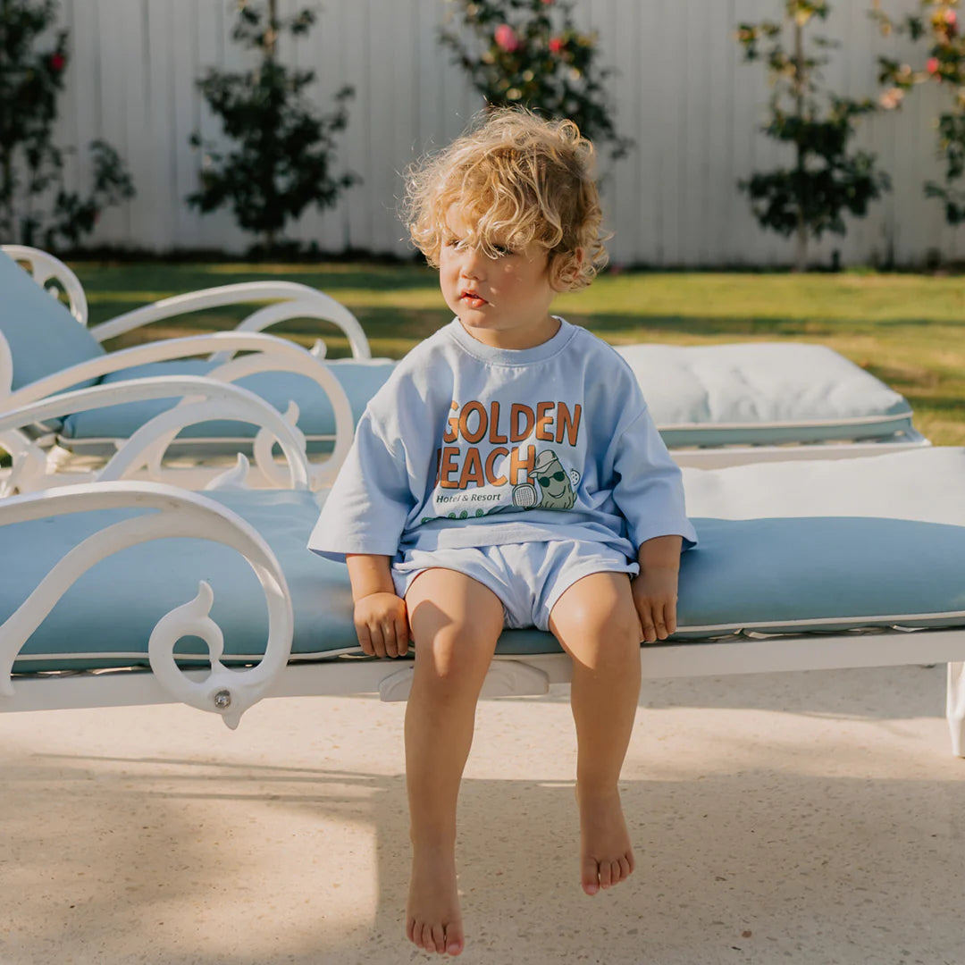 A young child with curly blonde hair sits on a light blue cushioned outdoor lounge chair, wearing a relaxed-fit "Golden Beach Mid Sleeve Tee" in Palm Blue by GOLDEN CHILDREN, alongside shorts. A white fence and greenery are in the background, suggesting a sunny day perfect for pickleball.