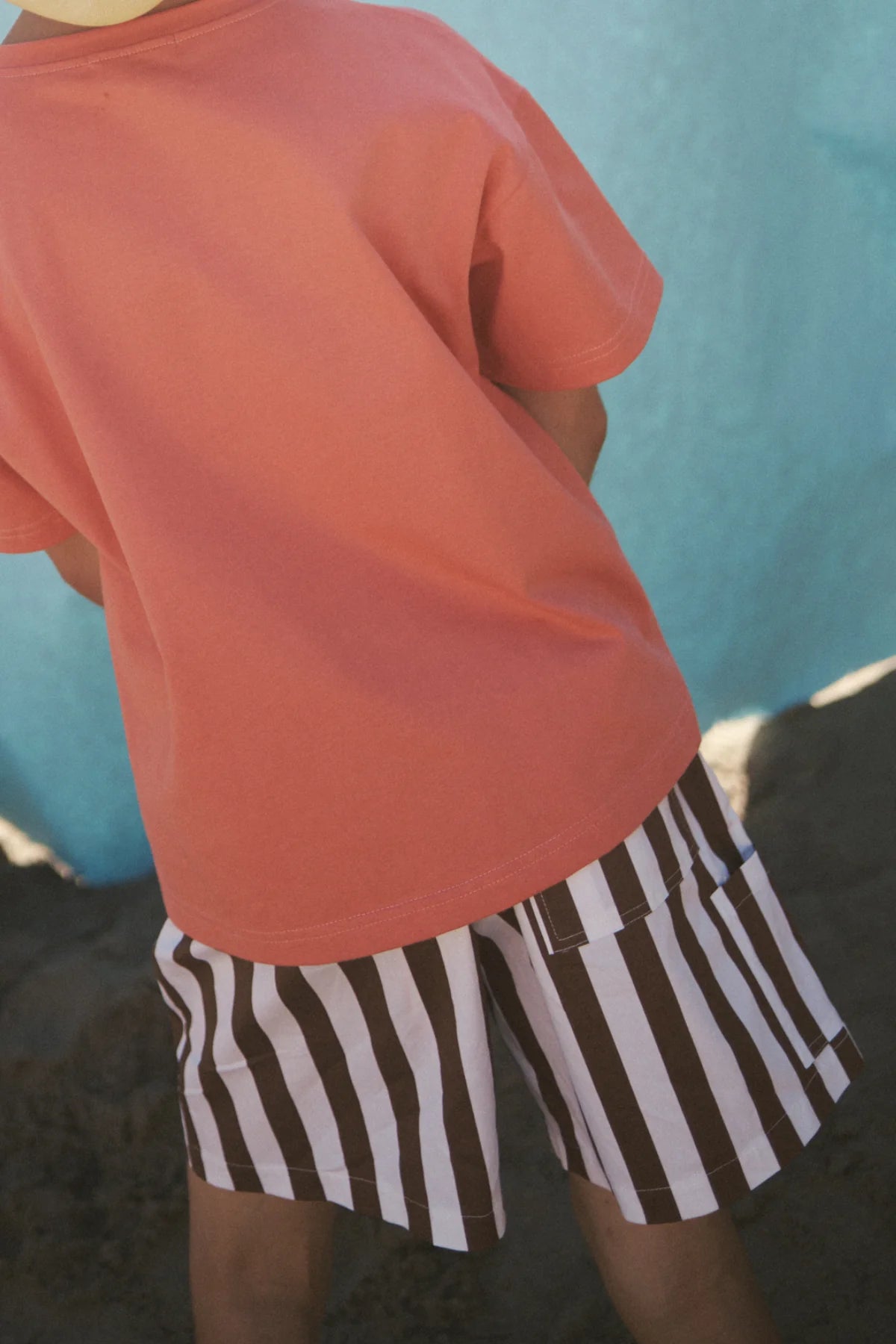 A child, wearing a coral t-shirt and LITTLE THE LABEL's Tei Shorts in brown/white stripe, stands on sand against a bright blue backdrop. The child faces away with no visible facial details.