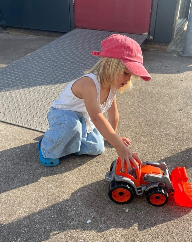 A child in a Ziggy Zaza "Roses Are Red" cap, white tank top, blue jeans, and blue shoes kneels on a concrete surface outdoors, playing with an orange toy vehicle next to a metal ramp. The cap features an adjustable brass clasp for the perfect fit.