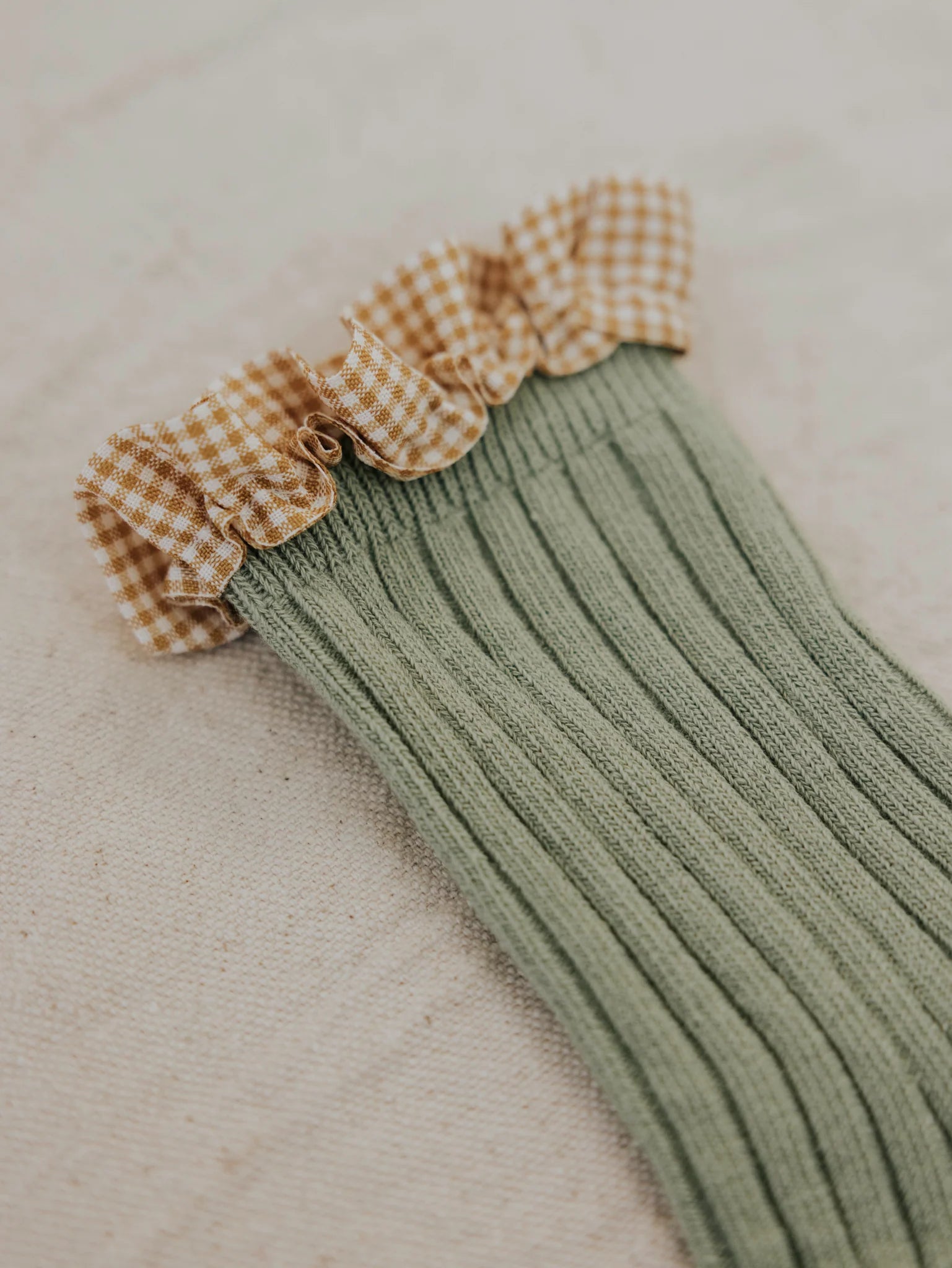 A single ribbed green Frilly Socks Avocado by VALENCIA BYRON BAY, featuring a ruffled gingham-patterned top in shades of brown and white, is displayed on a fabric background. The frilly socks' ruffles add a decorative touch to the cotton sock's design, making it perfect for any Valencia-inspired outfit.