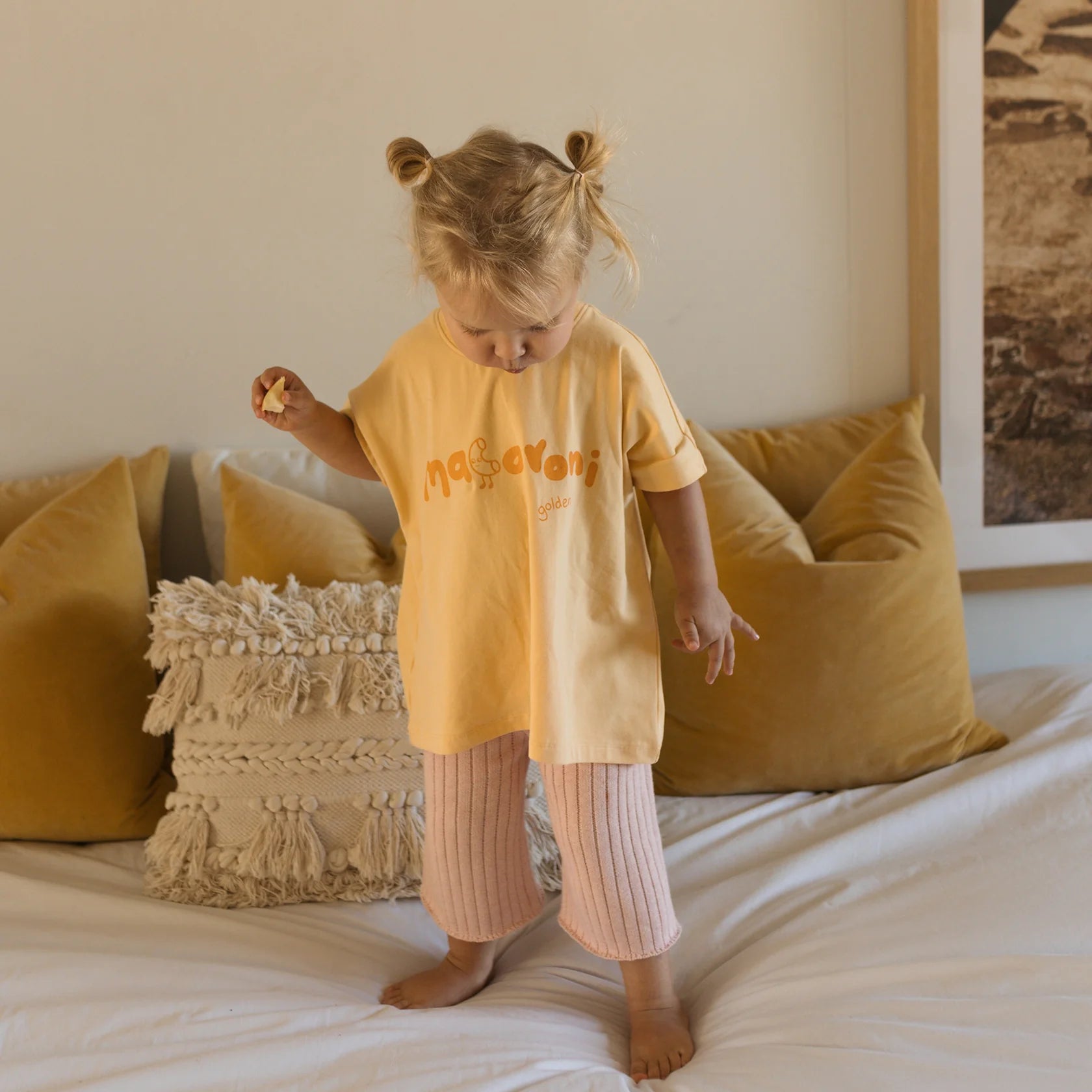 A young child with blonde hair styled in two buns stands on a bed. The child is wearing a light yellow "Macaroni Tee Golden Butter" from the brand GOLDEN CHILDREN, made of soft stretch fabric, along with light pink pants. The bed features mustard-yellow pillows and a textured cream pillow, complementing the room's neutral-toned décor.