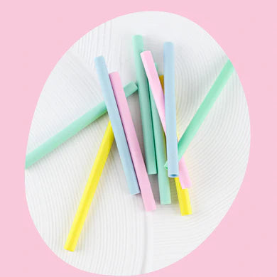 A cluster of colorful, eco-friendly silicone straws from the BBCINO Replacement Silicone Straw Set in pastel shades of yellow, green, blue, pink, and purple is spread out on a white, textured surface. The soft background is a light pink, creating a cheerful and clean composition.