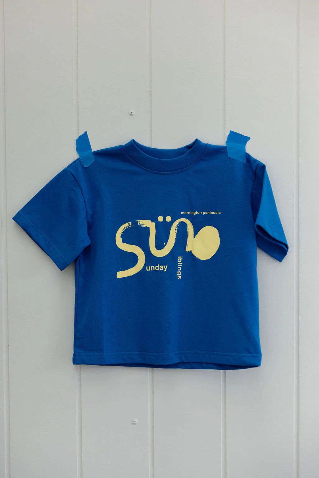 The Sunday Siblings ~ Il Sole Tee Blue/Yellow, a vibrant short-sleeved cotton shirt taped on a white wall, features an oversized fit for comfort, with "SÜ" and "Sunday Morning Brings" stylized graphics.