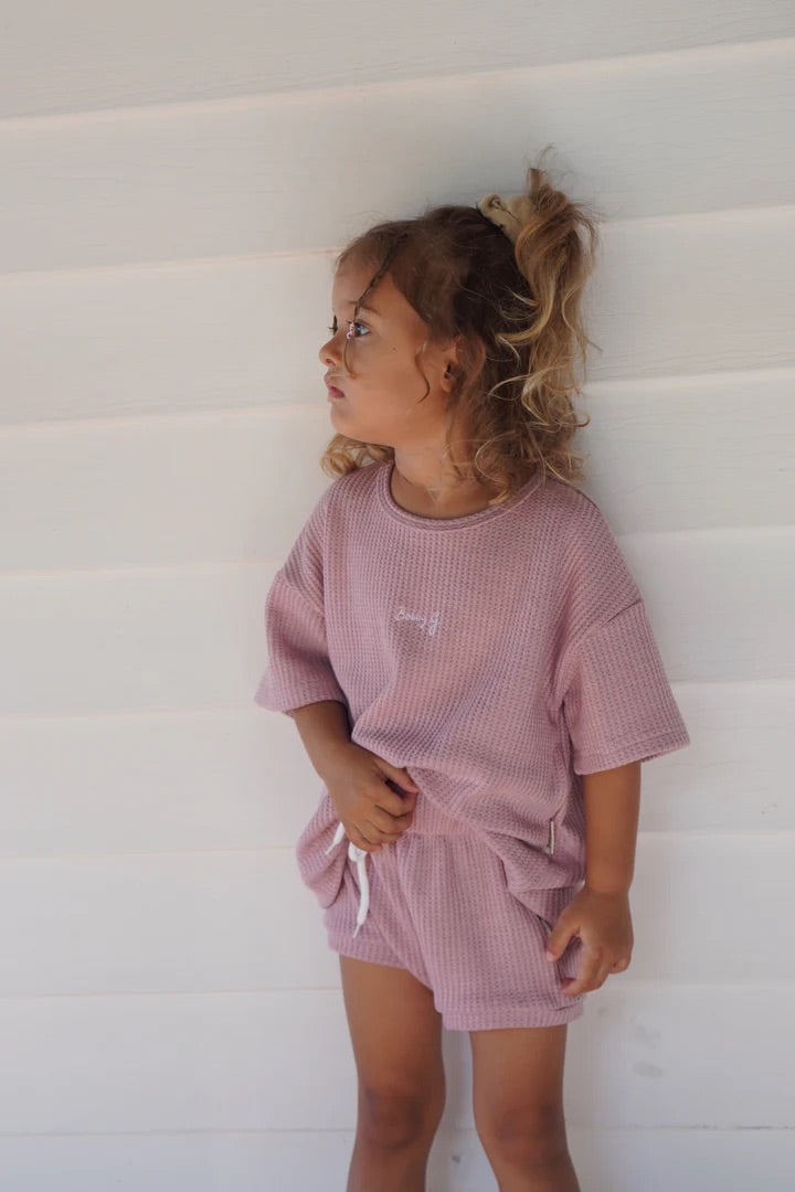 A young child with curly hair stands against a light wall, wearing the Coby Shorties Dusty Rose outfit by BOBBY G BABY WEAR. The soft waffle fabric ensemble includes a loose t-shirt and stretchy waistband shorts, gently lit by natural light.