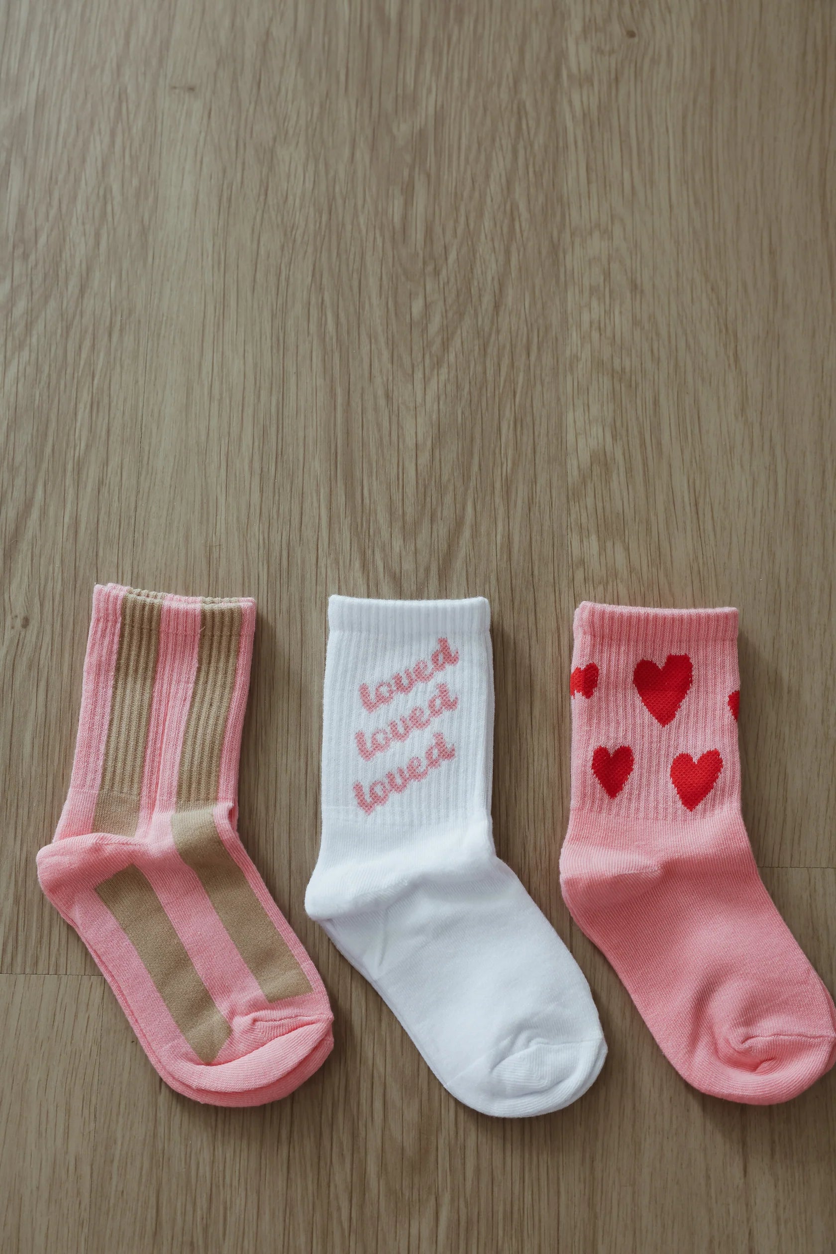 Three pairs of Circus Socks from TINY LOVE CLUB lie on a wooden floor, perfect for children ages 2-5 years. From left to right: pink and beige striped socks, white socks with "loved loved loved" written in pink, and pink socks adorned with red heart designs. All are machine washable for easy care.