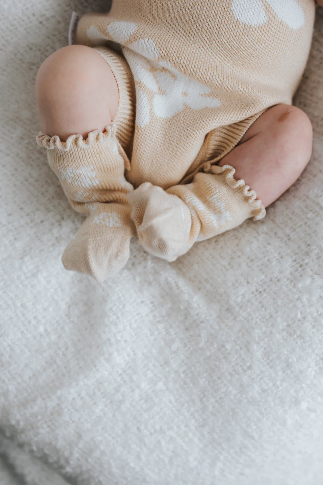 A close-up photograph of an infant's legs and feet wearing ZIGGY LOU's Socks Banksy. The baby is dressed in a soft beige outfit with white patterns, featuring cozy fabric and ruffled edges around the ankles—perfect for enjoying the Banksy + Beech Spring Collections. The baby rests on a textured white blanket.