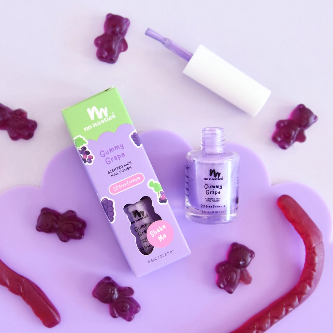 A bottle of NO NASTIES "No Nasties - Scented Scratch Off Kids Nail Polish Gummy Grape/Purple" with its box is displayed on a lavender cloud tray, surrounded by gummy candies. The scented polish brush rests on the tray.