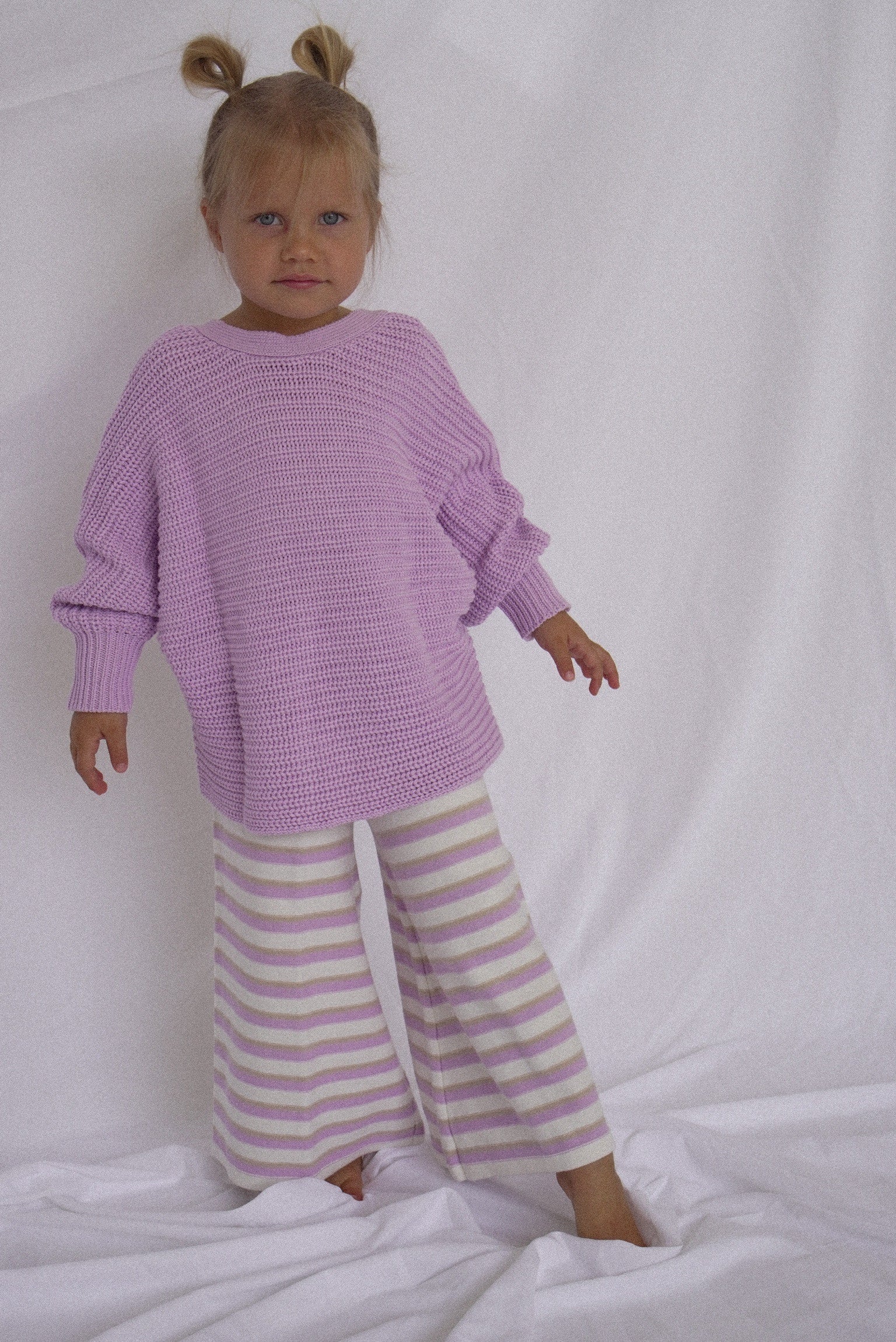 A young child with two buns stands against a white background wearing a lavender chunky knit sweater and En. The Label's Stripe Knit Pants in Lilac, featuring flared bottoms and crafted from cozy, stretchy fabric.