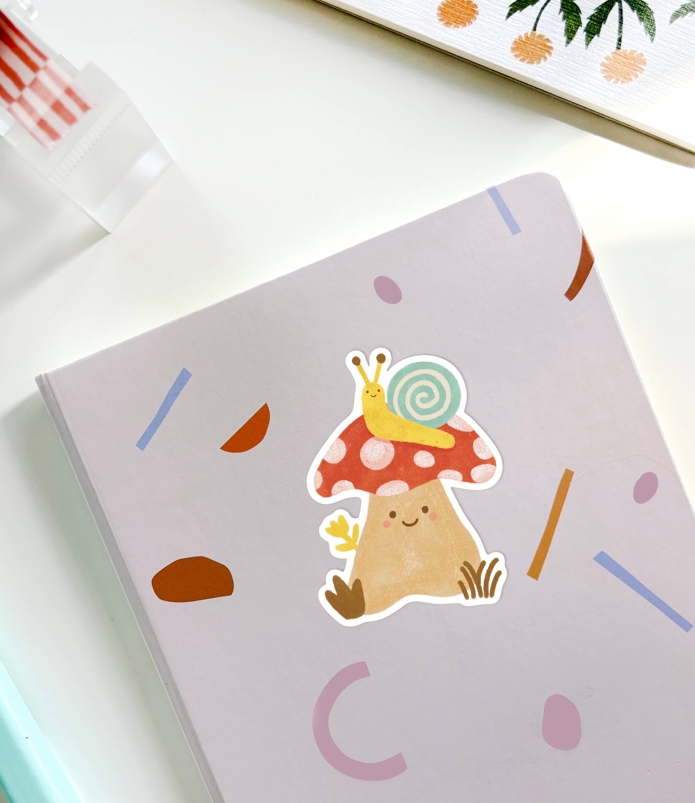 A close-up of a notebook featuring the whimsical Mushroom Sticker from LAUREN SISSONS STUDIO, which depicts a cute toadstool with a smiling face. A small snail with a spiral shell sits on top of the toadstool. The notebook cover is decorated with colorful, abstract shapes set against a white background.