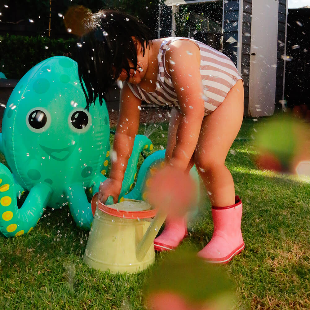 A child wearing Merry People ~ Andie Kids Gumboot Bubblegum boots and a striped swimsuit plays with a watering can on the grass. Water splashes around, and a large inflatable octopus is nearby as sunlight filters in, creating a lively scene.