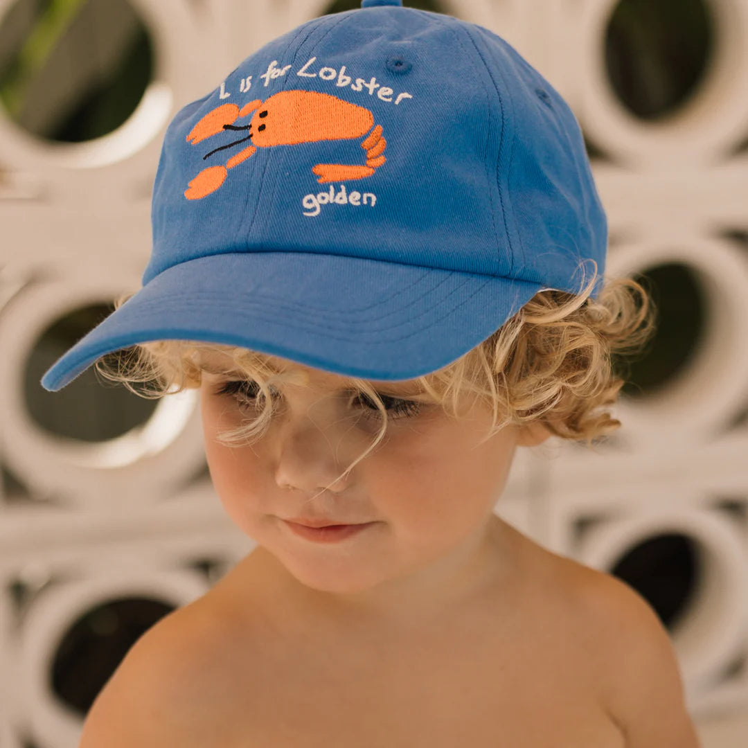 A young child with curly blond hair wears the Lobster Cap Deep Sea Blue, a blue dad cap designed with an orange lobster and emblazoned with "L is for Lobster" by GOLDEN CHILDREN. The adjustable brass clasp guarantees a perfect fit as the child stands against a wall with a white geometric pattern.