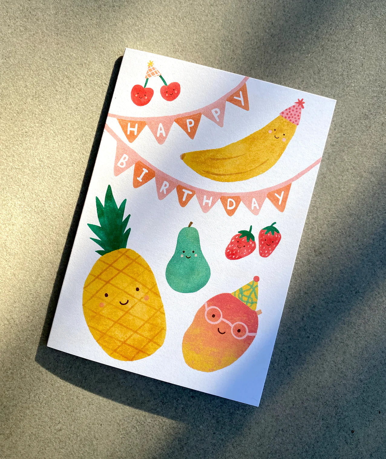 The "Birthday Greeting Card 'Fruit Party'" from LAUREN SISSONS STUDIO is an enchanting children's birthday card printed on recycled paper. It showcases delightful fruit characters, including a smiling pineapple, pear, cherry pair, banana, strawberries, and a bespectacled mango with a party hat. Above them, a banner joyously declares "Happy Birthday" against a light gray background.