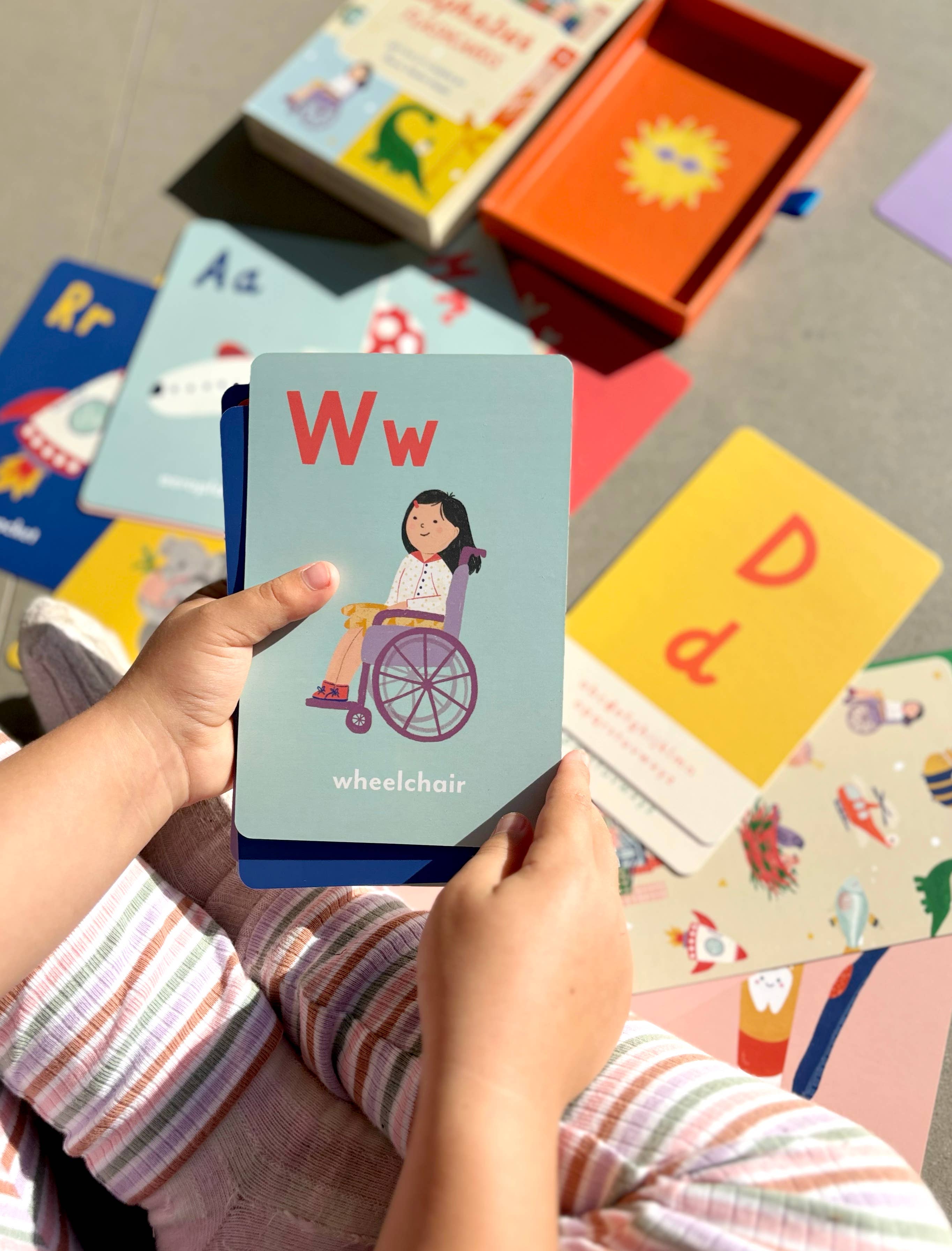 A child holds a laminated flashcard from the "A-Z Illustrated Alphabet Flashcards" by LAUREN SISSONS STUDIO, displaying the letter "W" with an illustration of a girl in a wheelchair and the word "wheelchair" beneath. Scattered on the floor in the background are additional alphabet flashcards and an orange box, part of their children's educational toys collection. The child is wearing striped pants.