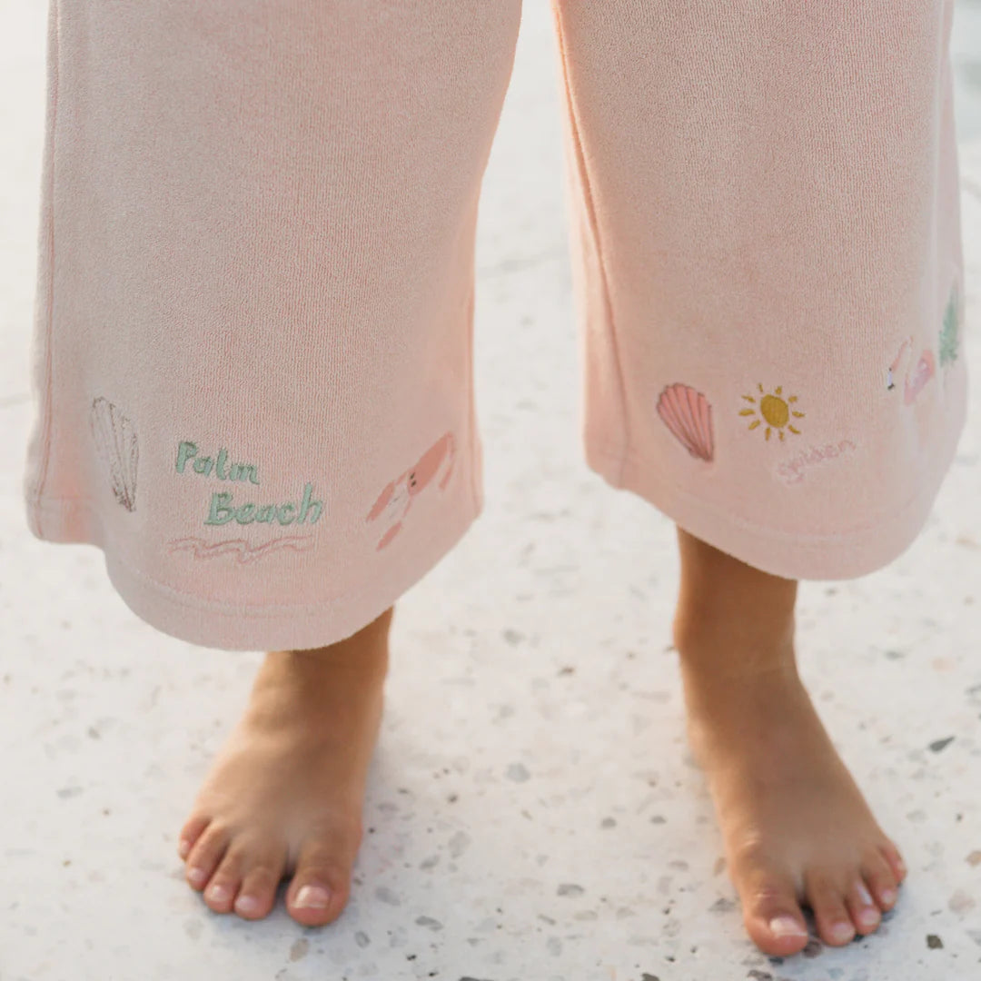 A child stands barefoot on a light-colored speckled floor, wearing Palm Beach Wide Leg Pants by GOLDEN CHILDREN. The pants are crafted from soft terry towel cotton in Flamingo Pink, featuring small illustrations like shells, a sun, and whimsical text.