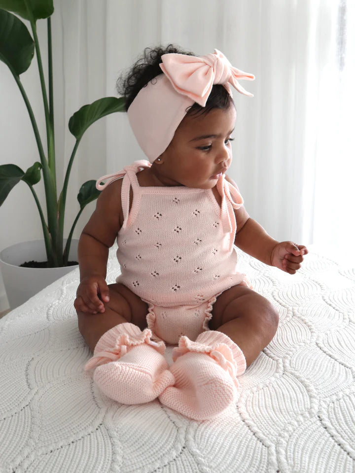 A baby sits on a white textured blanket, wearing a pink Frill Bodysuit Primrose from the ZIGGY LOU collection, matching socks, and a large bow headband. The baby is looking to the side with hands resting on the blanket. A green plant is visible in the background.