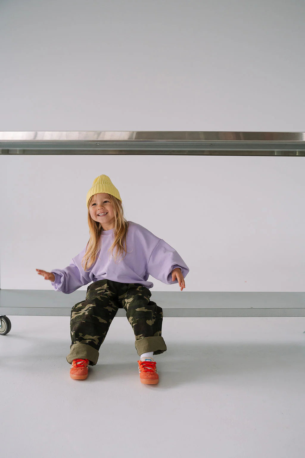 A smiling child in a yellow beanie, lavender sweater, and Sam the Label - The Cargo Pant Camo by SAM THE LABEL sits on the floor under a metal table with arms playfully outstretched against a plain white background.