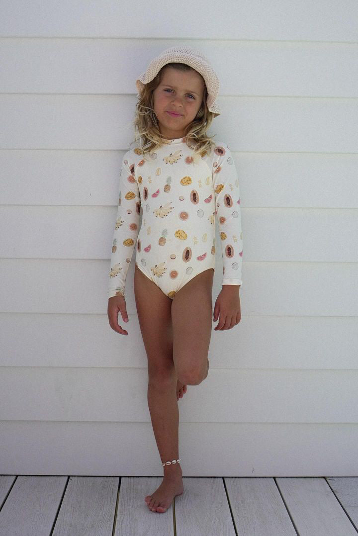 A young child wearing the EN. THE LABEL Tutti Frutti Long Sleeve One-Piece patterned with seashells and a white sunhat stands against a white wooden wall. They are resting one leg slightly bent and smiling, enjoying the day in their UFP 50+ rating, chlorine-resistant swimsuit from EN. THE LABEL.