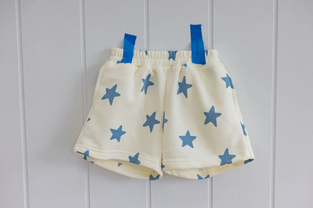 Sunday Siblings Kiddo Shorts Blue, made from cream french terry cotton with playful blue star patterns, feature an elastic waistband for comfort. Displayed on a white panel wall with blue clips, these stylish shorts ensure a trendy look.