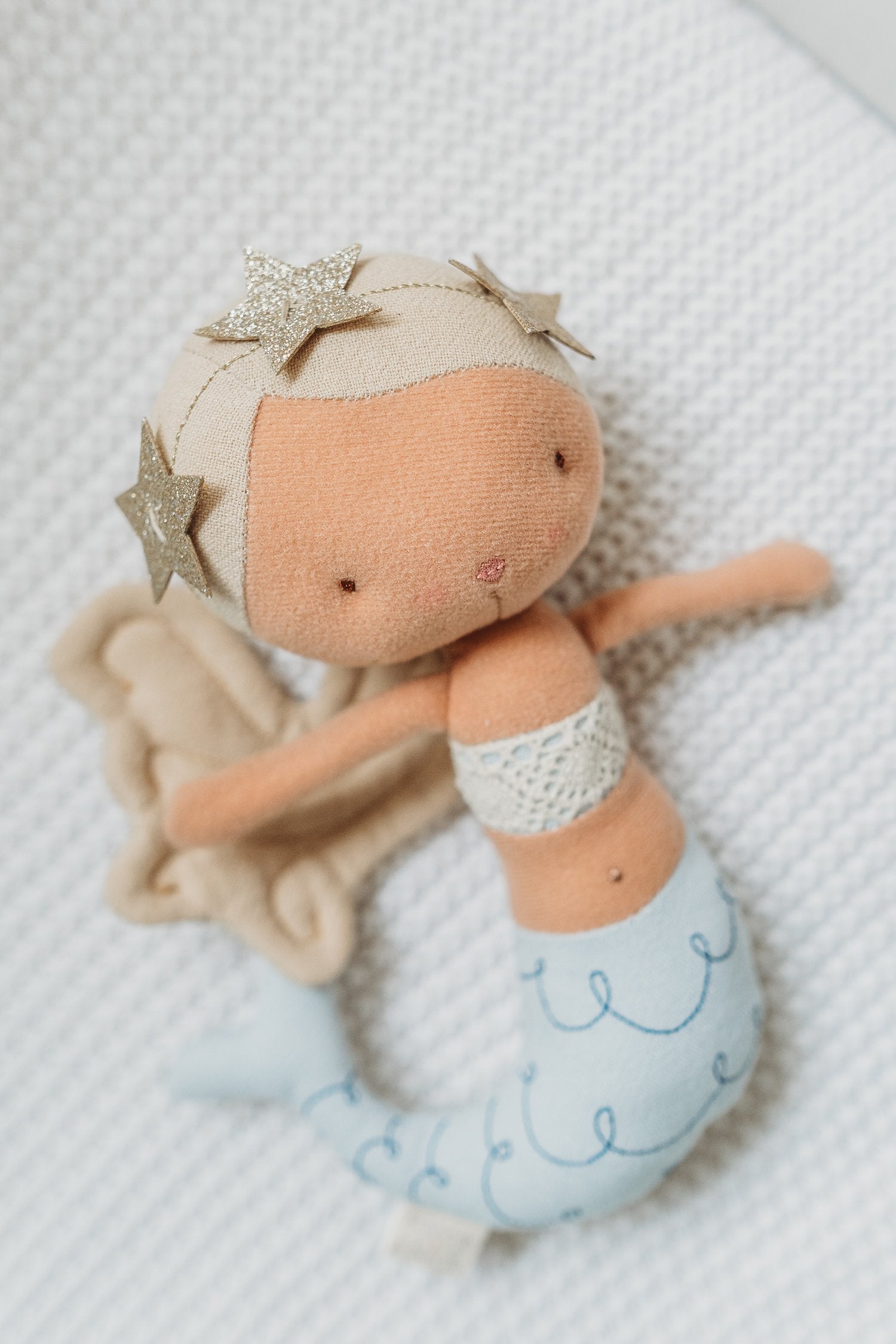 The Mermaid Pearl 22cm doll from PICCA LOULOU features a light brown body, blonde hair adorned with glittery starfish, and a blue tail. The doll is laying on a textured white surface, showcasing a small embroidered smile, simple facial features, and a white crochet top.