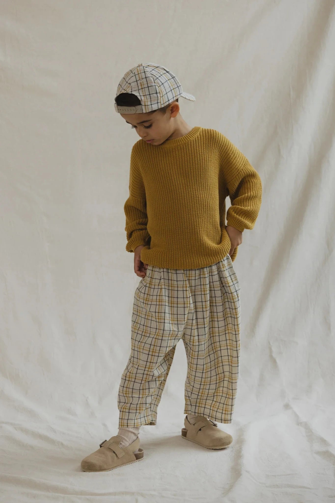 A young child stands confidently against a plain white backdrop, wearing unisex kids' clothing: a mustard yellow sweater, VALENCIA BYRON BAY's Rio Pant Picnic in 100% cotton plaid, and a matching plaid cap. The child also has beige shoes and strikes a casual pose with one hand resting on the hip and the other by the side.