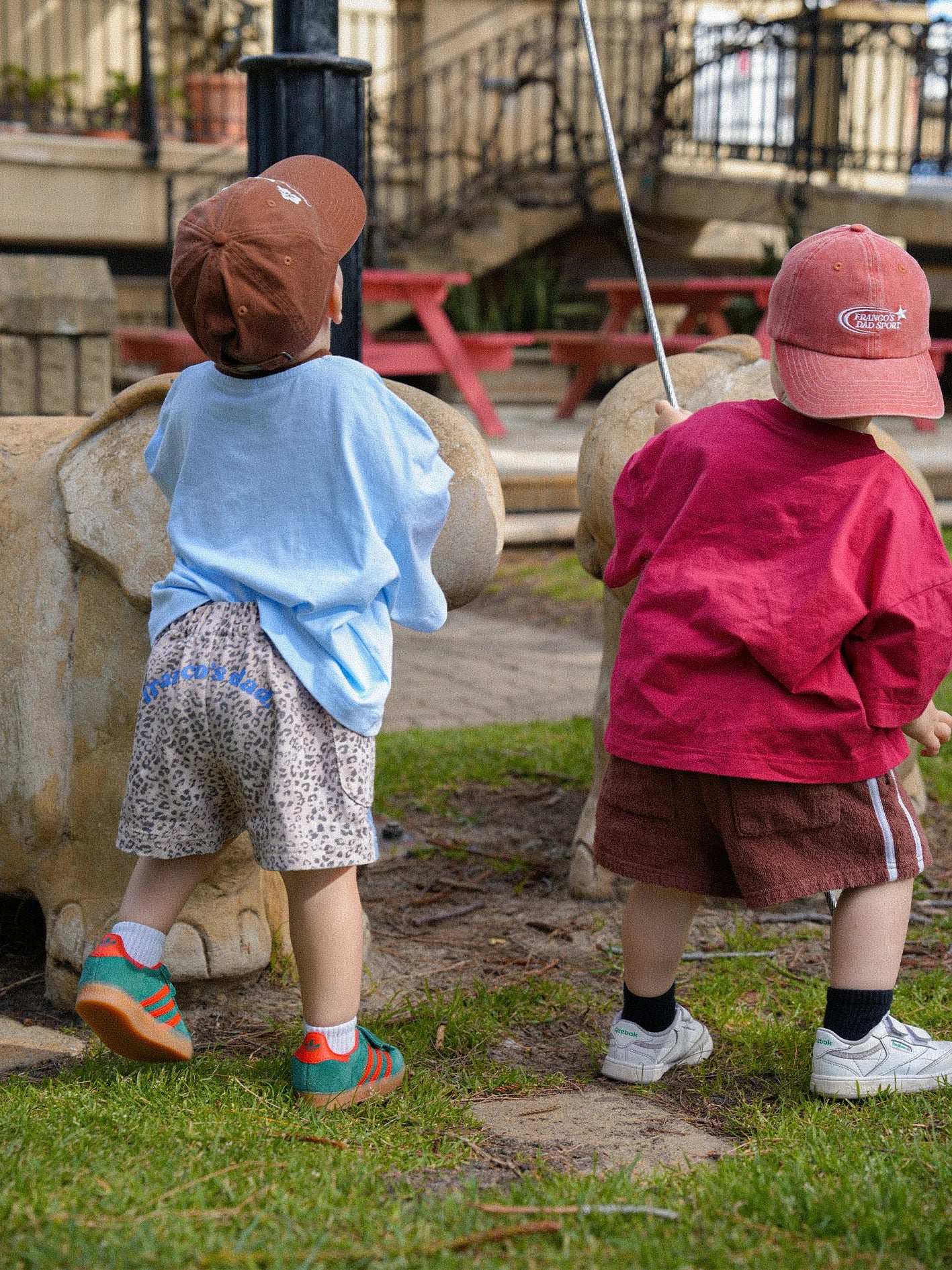 Two young children stand with their backs to the camera. One wears a blue shirt, leopard print shorts, a brown cap, and colorful socks; the other sports a red shirt and shorts topped with a PRE-ORDER Sports Cap Red by FRANCO'S DAD. They are outdoors on a grassy area near some playground equipment.