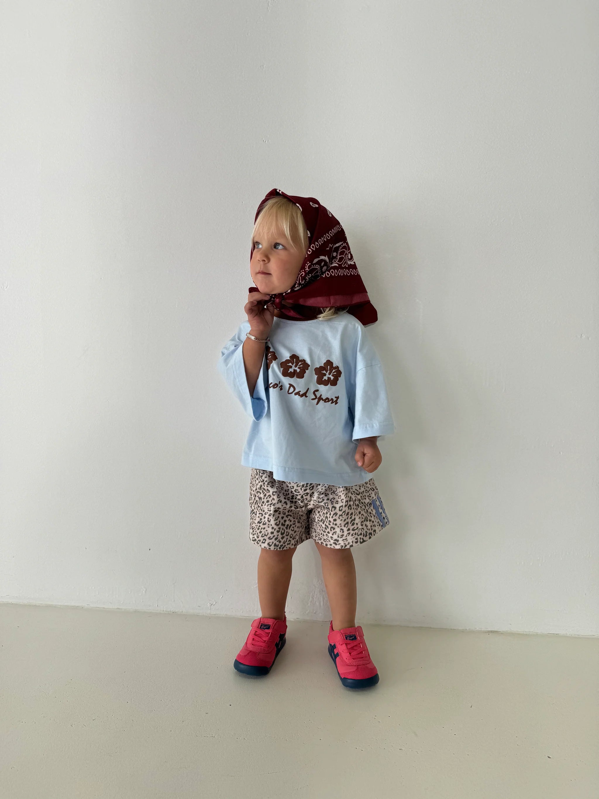 Against a plain backdrop, a toddler ponders thoughtfully while wearing an oversized light blue tee featuring text, patterned shorts, pink and blue sneakers, and a bandana adorned with a red hibiscus design. The tee is PRE-ORDER Hibiscus Tee by FRANCO'S DAD.