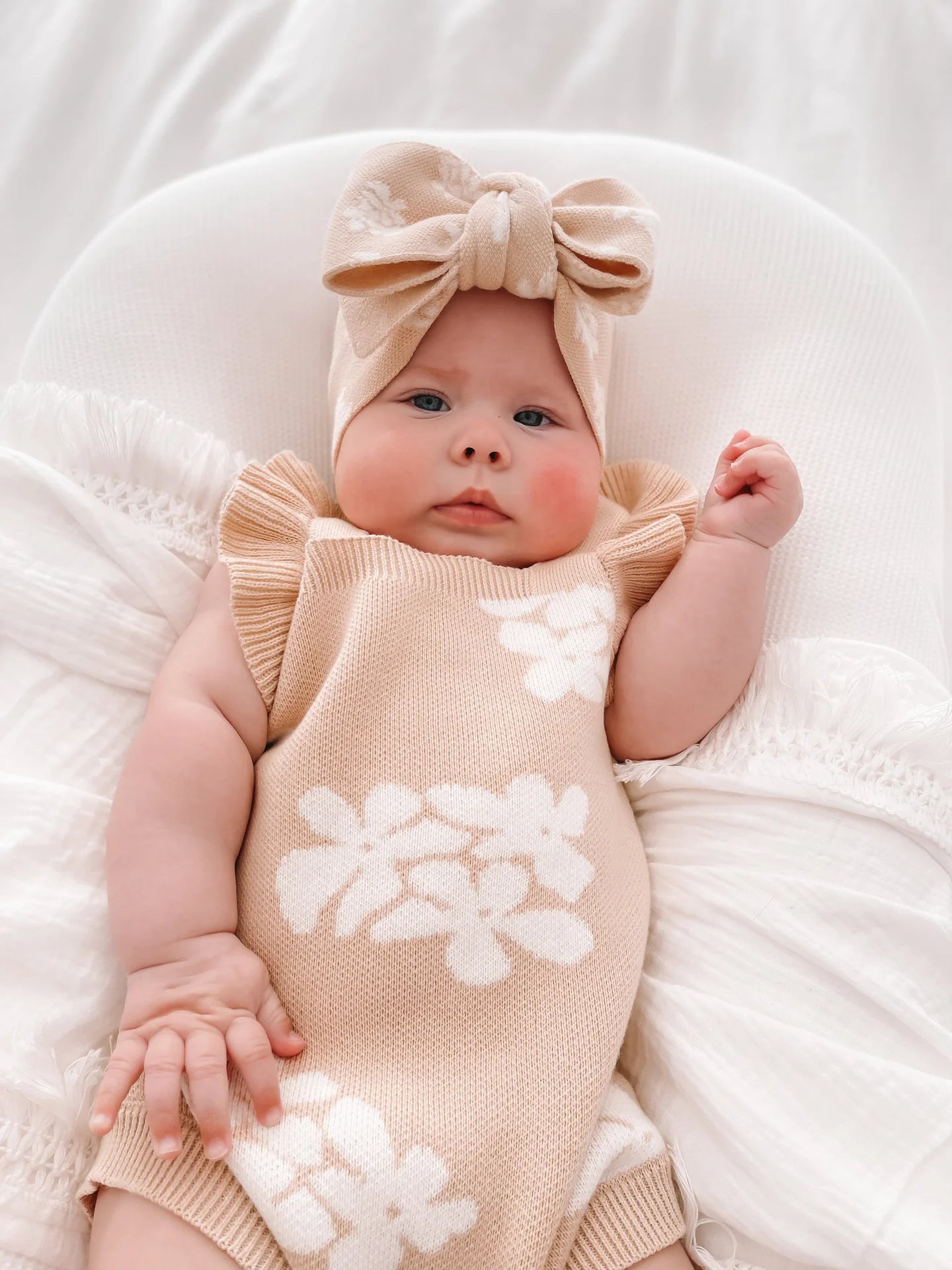 A baby lies on a white cushion wearing a ZIGGY LOU Frill Romper Banksy, a beige knit outfit adorned with white floral patterns, complemented by a matching beige headband with a large bow. The romper features functional snaps for easy changing. One of the baby's hands rests on the cushion, while the other is slightly raised, giving an appearance of calm and contentment.