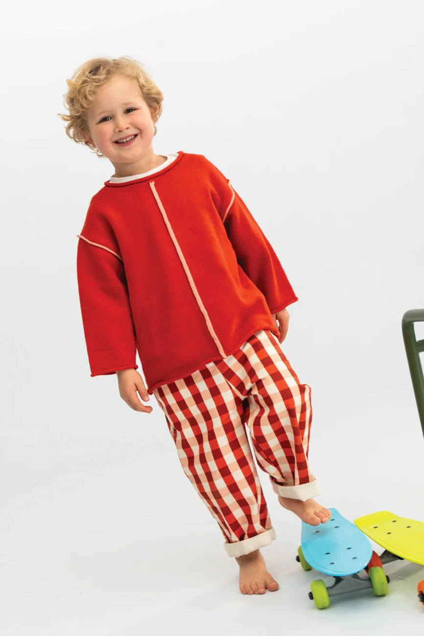 A young child with curly blonde hair and a happy expression is wearing a red long-sleeve shirt and the Picnic Pocket Pants Cherry by SONNY LABEL, which feature a red-and-white picnic pattern made of 100% cotton. The child is standing on one foot while the other foot rests on a green and blue skateboard, against a plain white background.