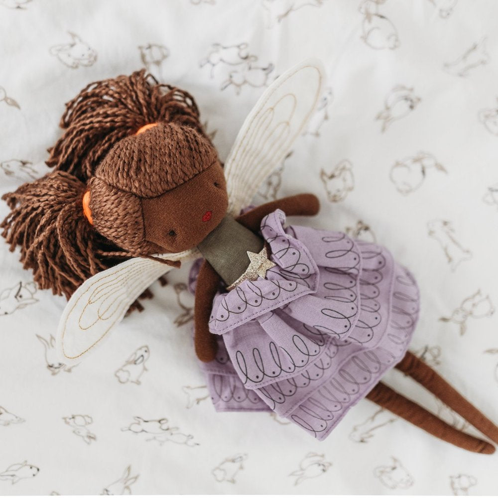 A delightful handmade Fairy Feline 35cm doll from PICCA LOULOU, featuring brown yarn hair, wings, and a lavender dress adorned with a small star on the chest, lies on a white sheet patterned with small bunny illustrations. This enchanting piece brings whimsical charm to any room.