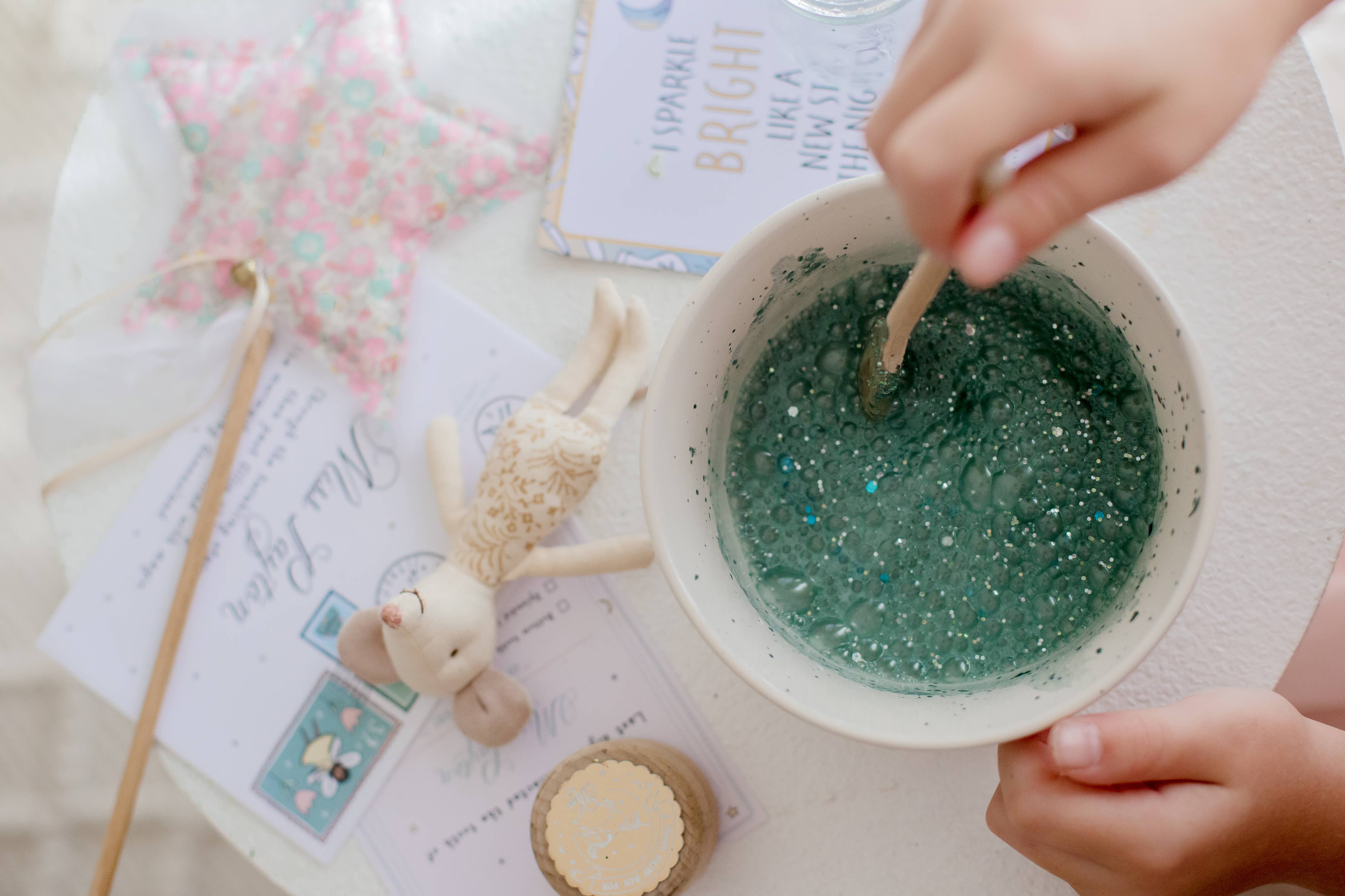 A child stirs a teal, glittery concoction from the Tooth Fairy Magic Potion Pouch by THE LITTLE POTION CO in a bowl, creating a mystical elixir. Nearby lie a floral star wand, a small cloth doll, and an illustrated card. Books filled with whimsical themes and sparkling ingredients are spread across the white surface.