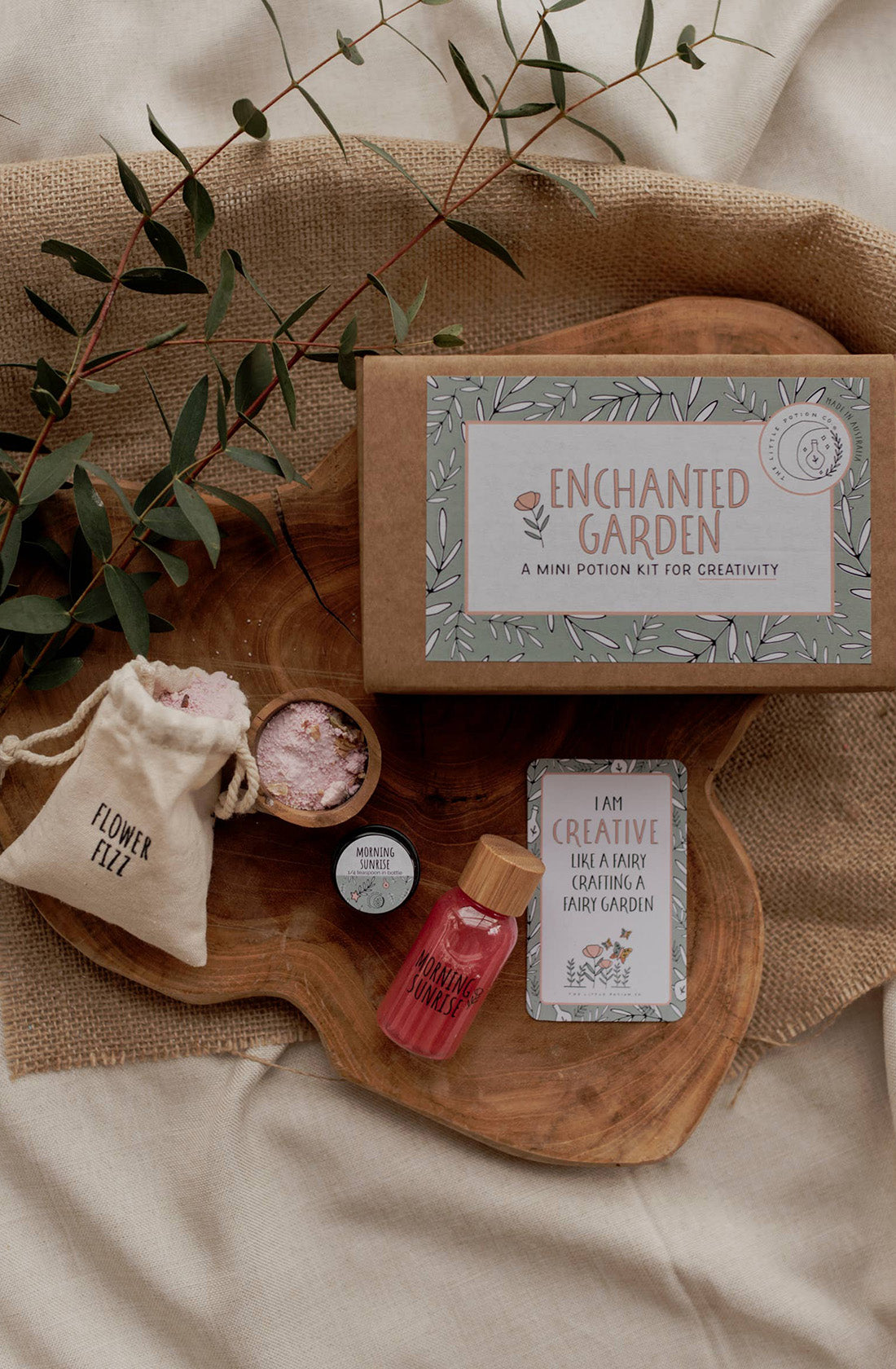 A flat lay of the "MINI Enchanted Garden Potion Kit (A potion for creativity)" by THE LITTLE POTION CO., artfully displayed on a rustic wooden surface with a beige cloth underneath. Ideal for sensory play, the kit includes a flower fizz pouch, a small bottle of botanical magic powder, a vial of pink liquid, and a creativity card. Sparse greenery decorates the setup.