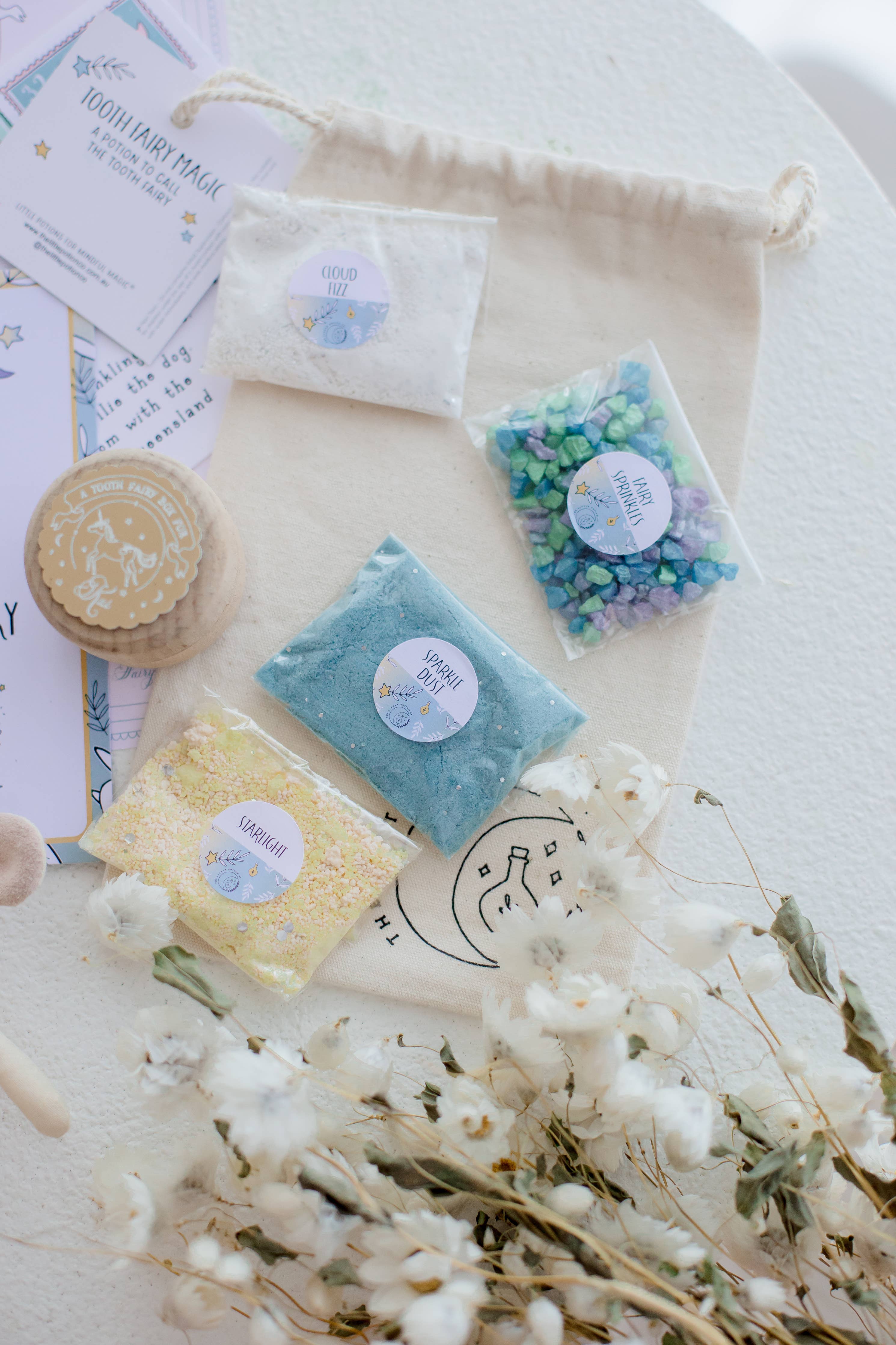 A flat lay of craft supplies on a table, featuring pastel-colored stones resembling sparkly ingredients in small bags, the Tooth Fairy Magic Potion Pouch by THE LITTLE POTION CO, floral decorations, and paper materials.