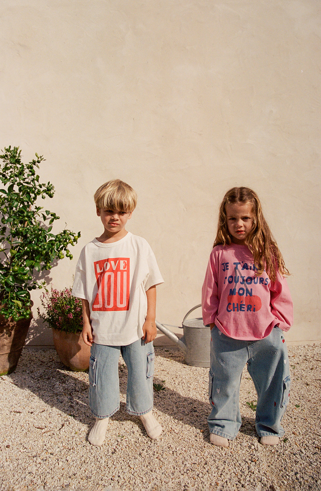 Two children stand outside on a gravel surface. The child on the left, wearing an oversized fit white "Je T’aime Long Sleeve Tee Mini" by THE WHOLESOME STORE with "LOVE YOU" in red letters and blue jeans, has blond hair. The child on the right, with long brown hair, sports a pink shirt and blue jeans. A plant and watering can are in the background.