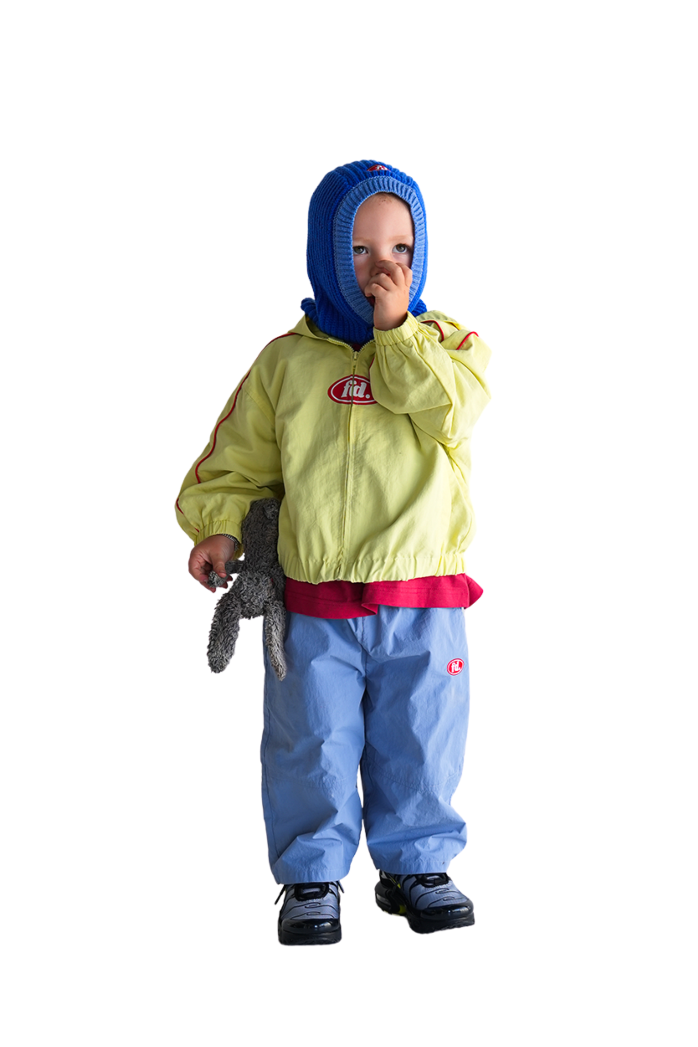 A young child wears an oversized Franco's Dad ~ Tech Spray Jacket Butter by FRANCO'S DAD, with blue pants and a hood. They clutch a stuffed animal in one hand, the other near their mouth, against a white background.