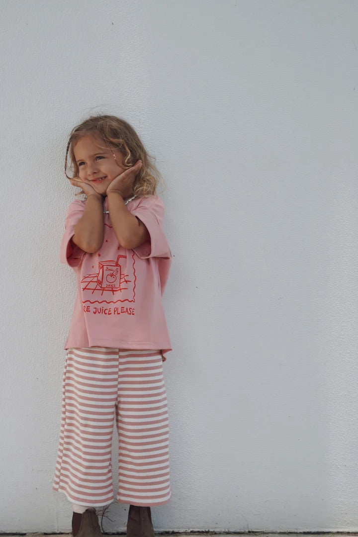 A young child with long hair smiles with hands on their cheeks, wearing BOBBY G BABY WEAR’s Candy Cane Pink Floaty Pants and a pink T-shirt featuring a juice box graphic and "More Juice Please" text, standing against a plain white wall.