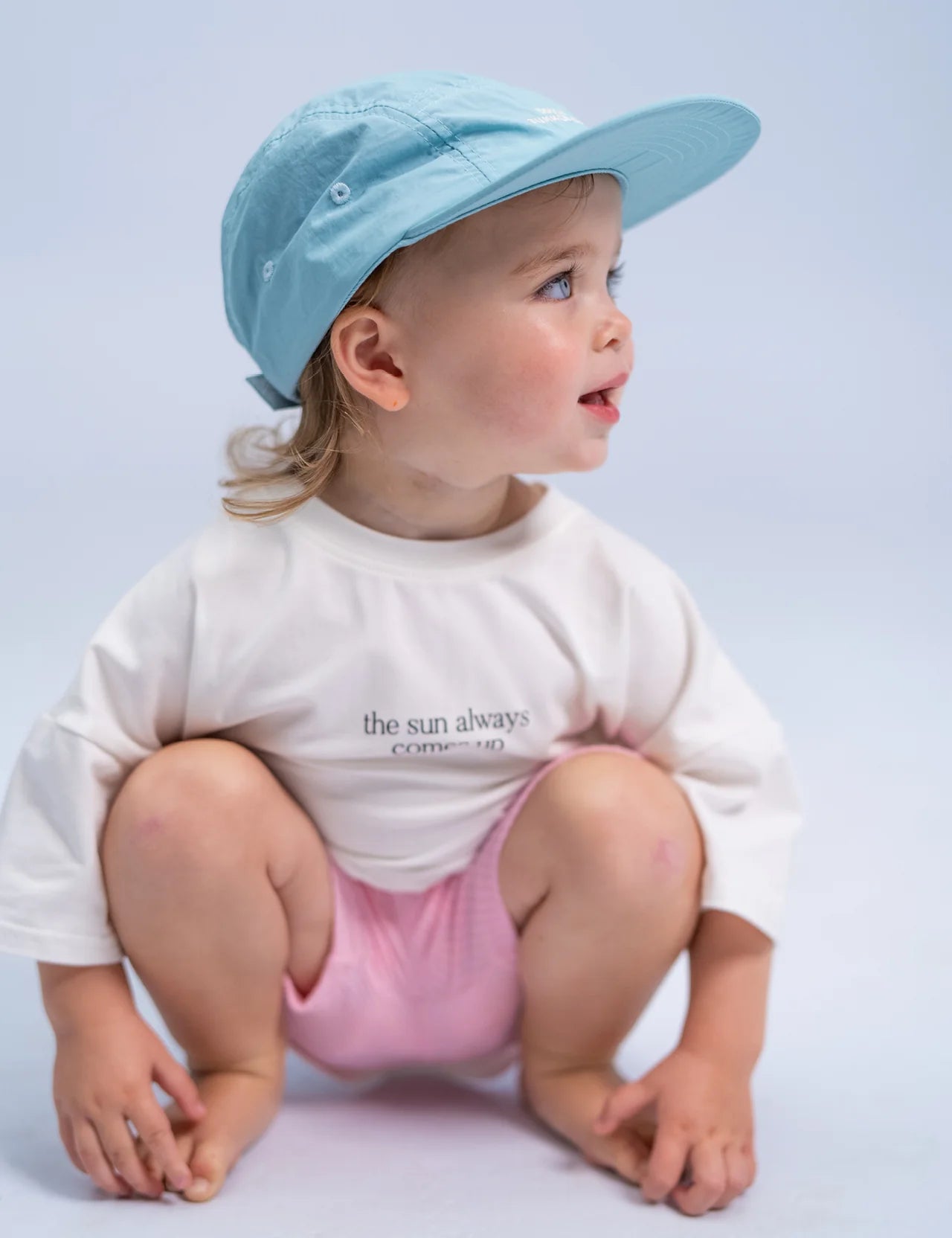 A toddler wearing the SONNY LABEL Summer Club Cap in Ocean, featuring an adjustable strap, and a white shirt with the message "the sun always comes up," crouches while looking to the side. They are styled with pink shorts against a light blue background.