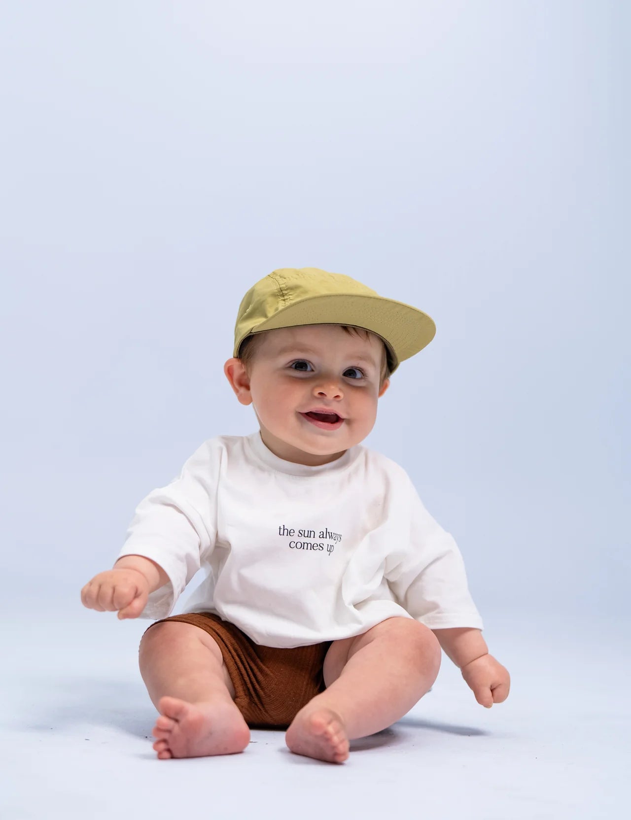 A smiling baby wearing the SONNY LABEL Summer Club Cap in sage, featuring an adjustable velcro strap, sits on a light blue background. The baby is dressed in a white shirt with the text "the sun always comes up" and snug brown shorts, with bare feet adding to the charm.