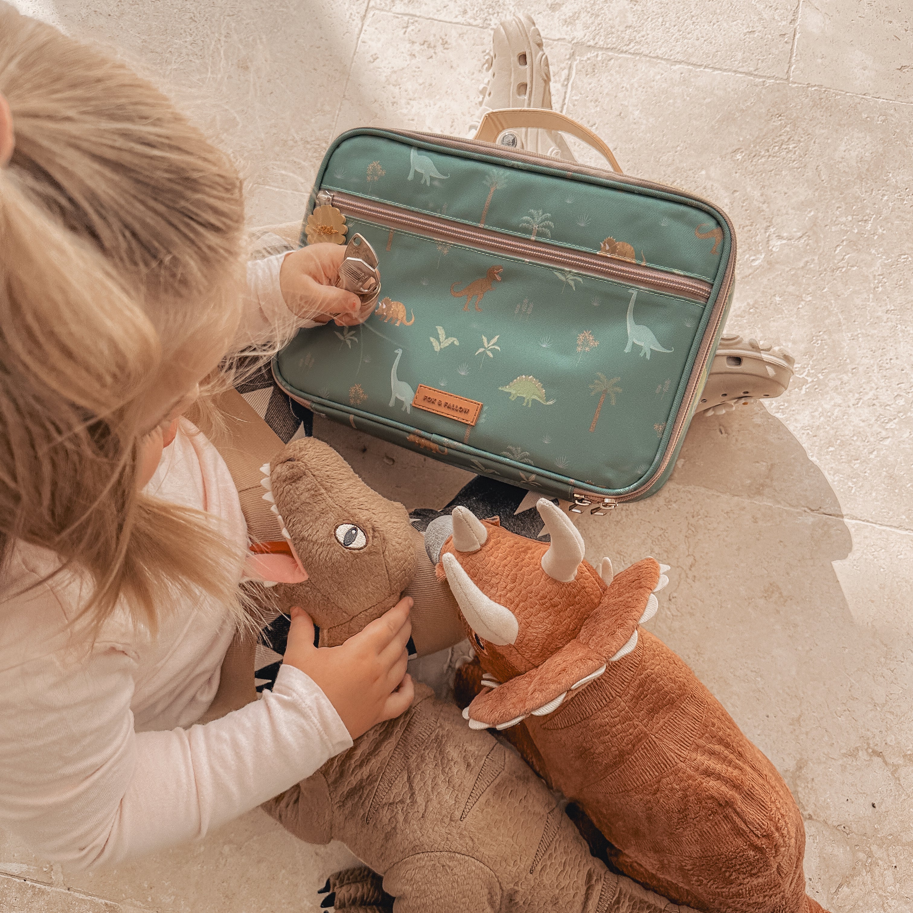 A child is sitting on a light-colored floor holding two plush dinosaur toys, a T-Rex and a Triceratops. In front of the child is a green dinosaur-themed backpack from FOX & FALLOW, featuring various dinosaur illustrations and their Dinos Lunch Bag. The scene is bathed in warm, natural light.