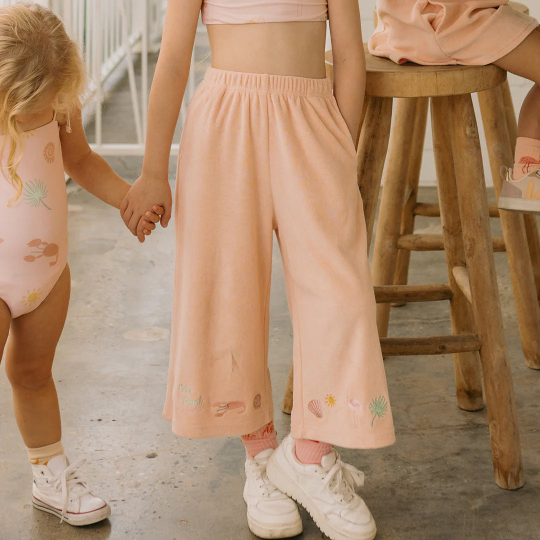 Two young children hold hands, one wearing the playful Palm Beach Wide Leg Pants in Flamingo Pink and the other sporting a matching sleeveless top. They enjoy a relaxed indoor setting with a wooden stool nearby, their charming outfits from GOLDEN CHILDREN adding to the scene's cozy appeal.