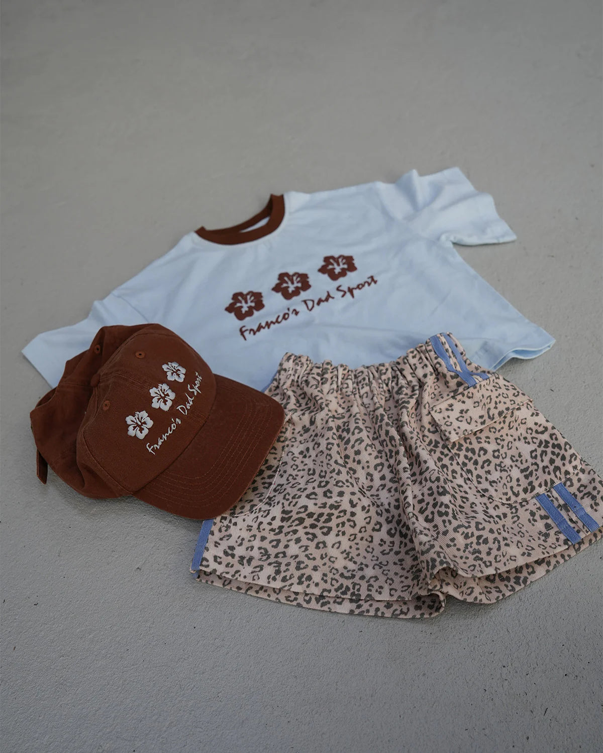 A laid-out outfit features the PRE-ORDER Hibiscus Tee by FRANCO'S DAD, an oversized light blue t-shirt with brown lettering and logos, paired with beige leopard print shorts and a brown baseball cap that sports matching logos and text. Capturing a hint of 90s sports design, the items are elegantly displayed on a concrete surface.