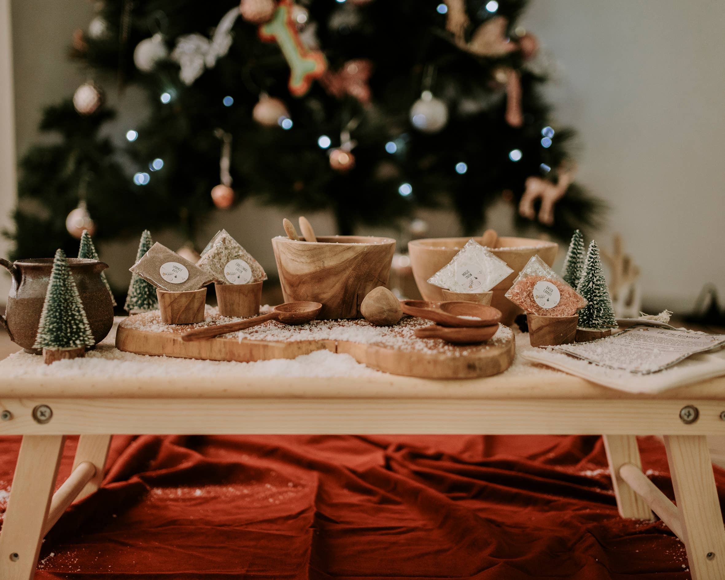 A festive table setting showcases wooden bowls and small trees embellished with ornaments on a wooden tray. A sprinkle of fake snow whimsically decorates the scene. In the blurred background, a Christmas tree glows with lights and ornaments, creating an enchanting ambiance similar to a magical tradition. To add a touch of holiday magic, consider including THE LITTLE POTION CO's Xmas Eve Potion Pouch to Call the Reindeer as part of your setting.