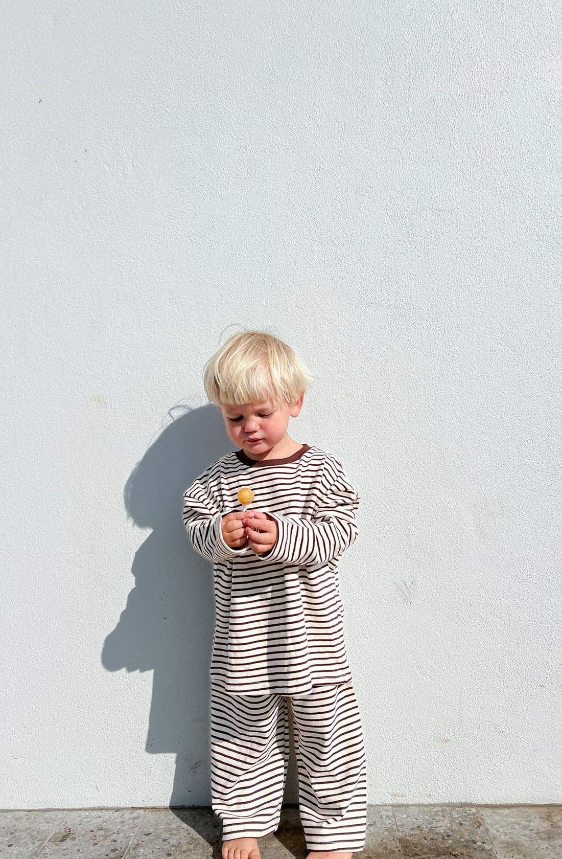 A young child with blond hair stands barefoot against a plain white wall, wearing the Every Day Set Choc Chip pajamas from LENN LABEL, made of 100% knit cotton. Featuring an elastic waistband for added comfort and an oversized fit, the striped pajamas add to the cozy look. Holding a small toy with both hands, the child looks down at it intently while a shadow is cast on the wall behind them.