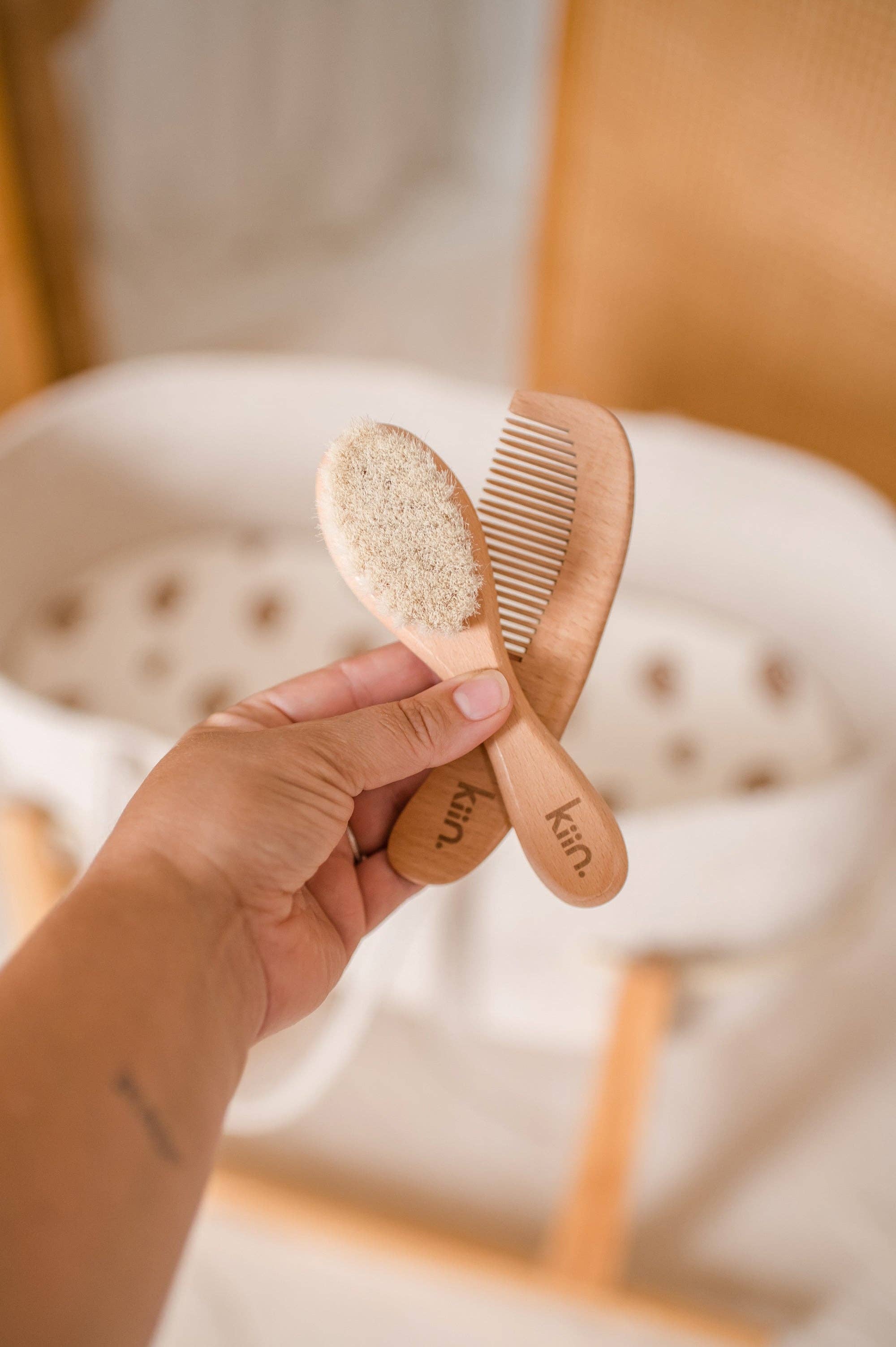 A hand grasps the KIIN BABY Wooden Baby Brush and Comb Set, featuring eco-friendly goat wool bristles. The backdrop showcases a softly blurred, cozy cradle design.