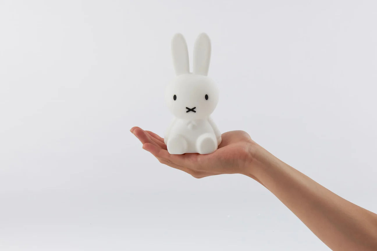 In the palm of a person's extended hand sits a small, white, rabbit-like figure with long ears. This is the Bundle of Light - Miffy by MR MARIA, a soft silicone LED lamp that adds charm to any space with its gentle glow. The plain white background enhances its minimalist appeal, making it perfect as a gift display item.