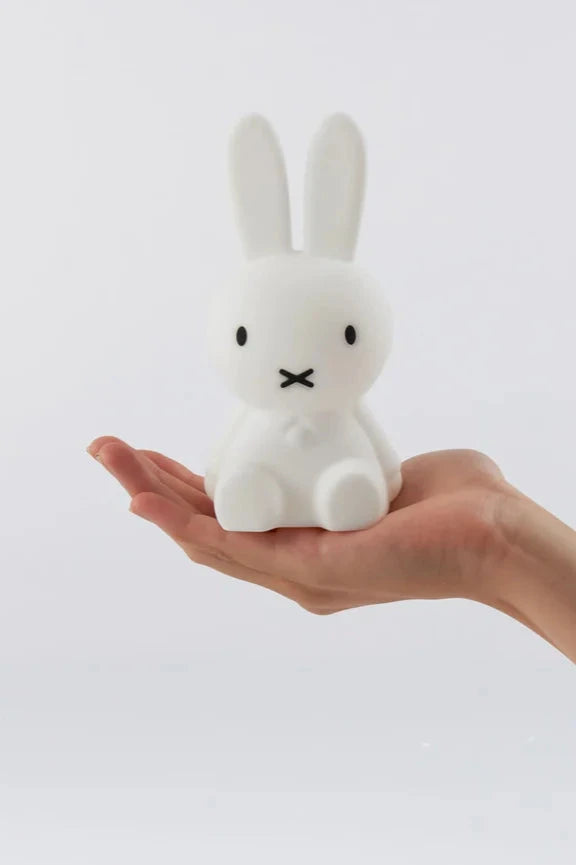 In the palm of a person's extended hand sits a small, white, rabbit-like figure with long ears. This is the Bundle of Light - Miffy by MR MARIA, a soft silicone LED lamp that adds charm to any space with its gentle glow. The plain white background enhances its minimalist appeal, making it perfect as a gift display item.