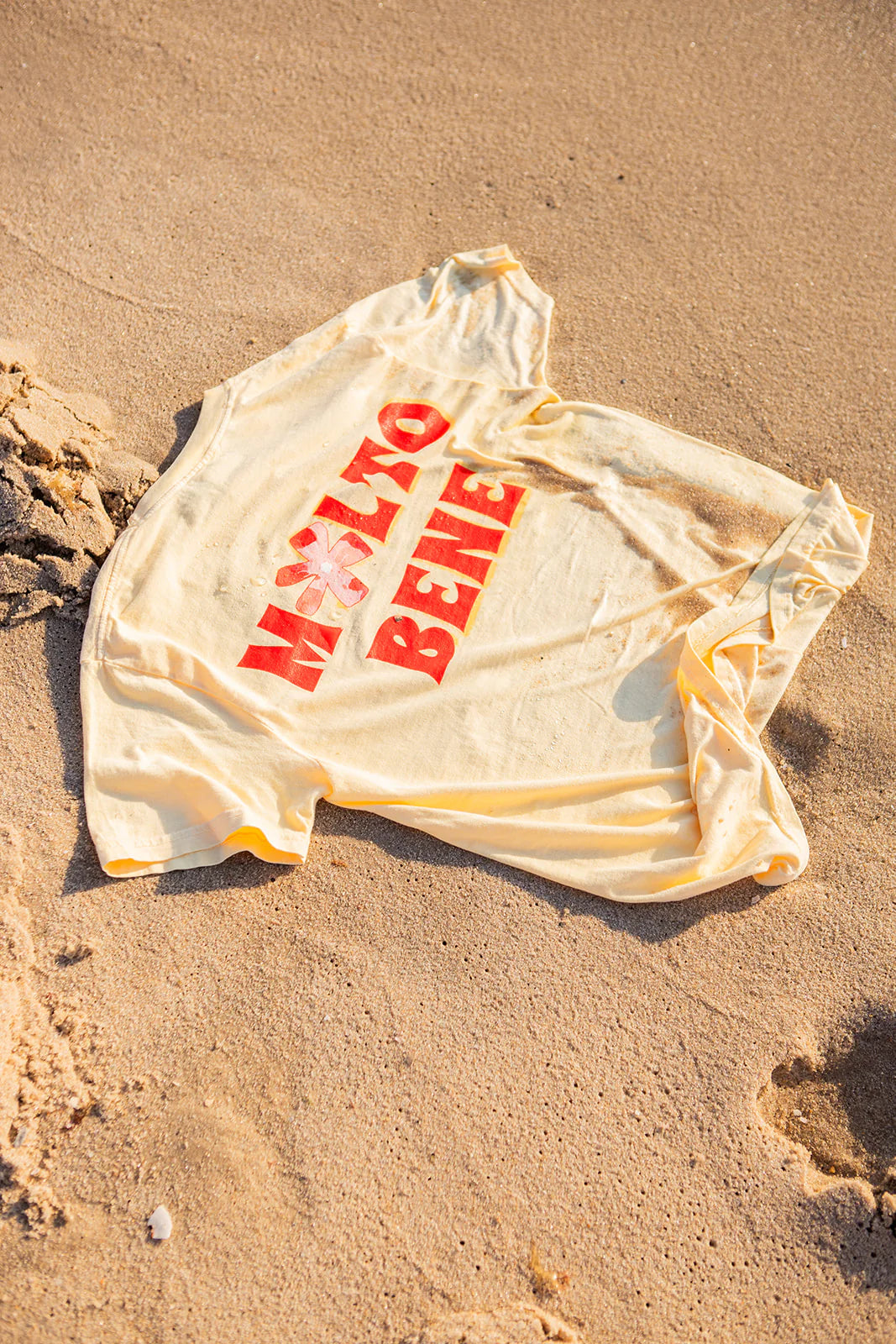 The Molto Bene T-Shirt by MAKU THE LABEL is a boxy crop tee made of 100% cotton, featuring "Molto Bene" and a hand-painted vibrant red flower elegantly resting on the sandy beach.